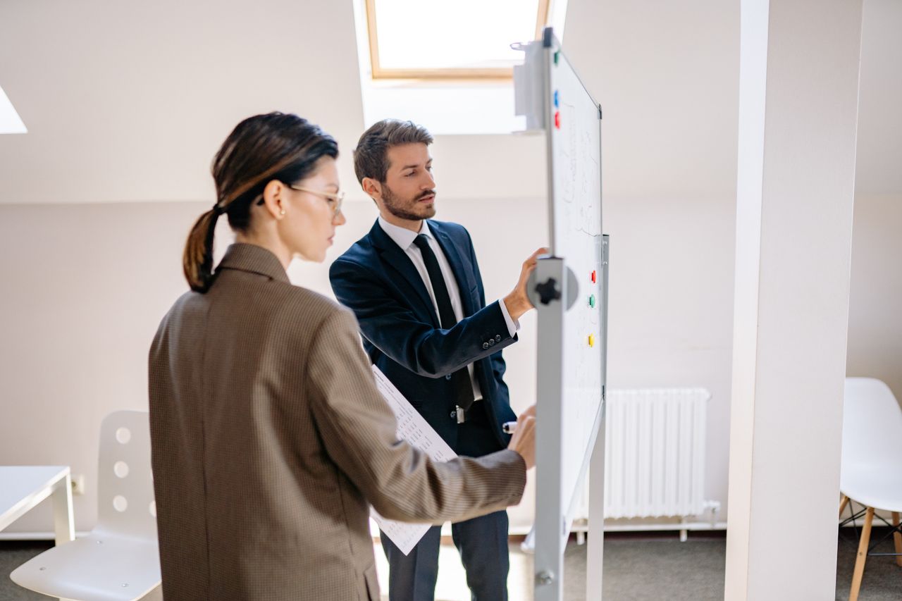 Two colleagues in formal attire discussing strategies on a whiteboard in a modern office space.